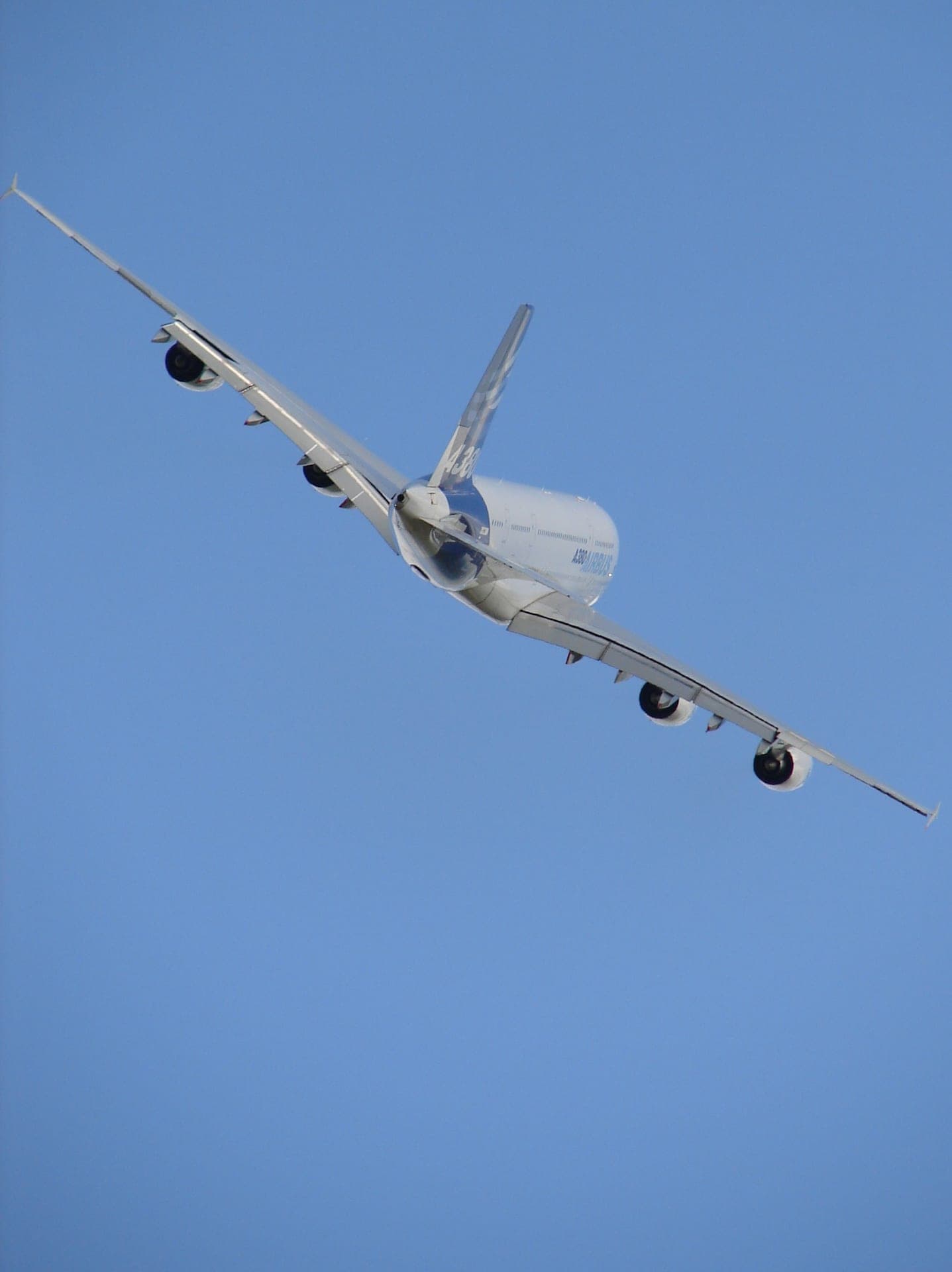 An Airbus A380 banking steeply, its immense wing planform and four engines seen from below against the sky