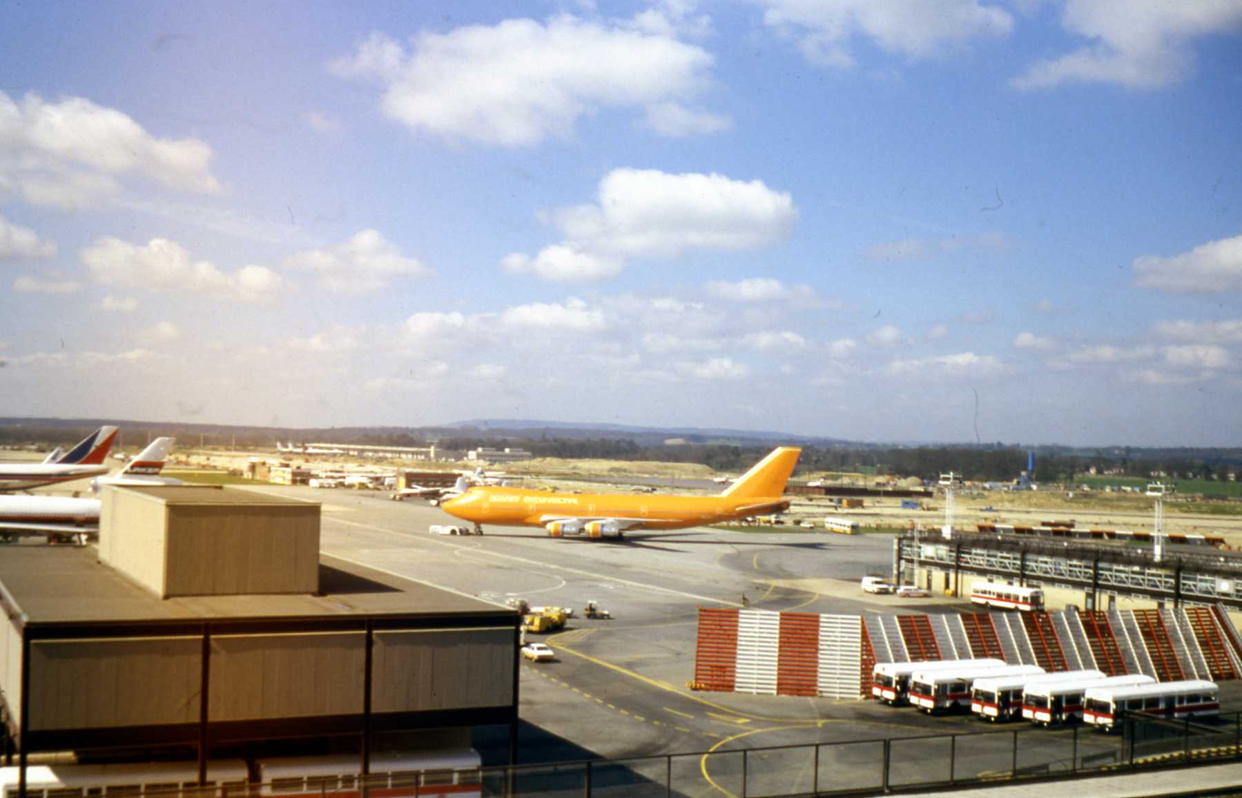 An orange-liveried Boeing 747-200 freighter taxiing past a terminal building in the late 1970s or early 1980s