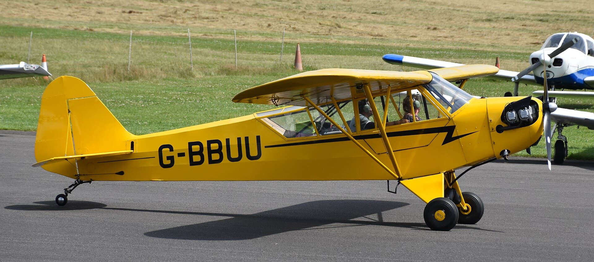 A yellow Piper J-3 Cub, registration G-BBUU, parked on an airfield apron