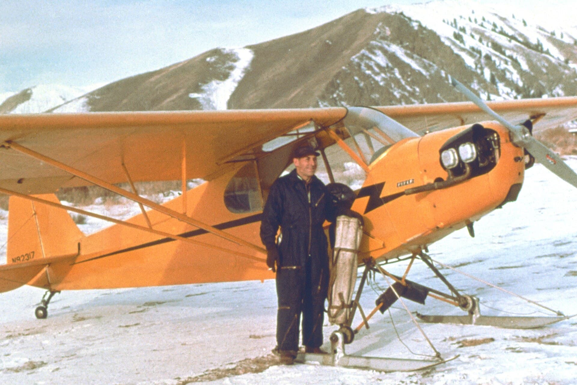 An orange Piper Cub fitted with skis, parked on snow in a mountain valley