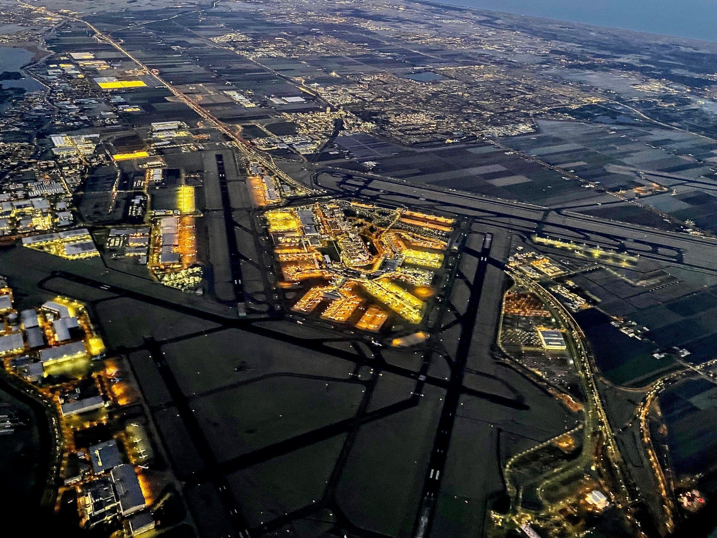 Schiphol at dawn from an aircraft climbing away: the lit terminal at the centre of its six runways, spread across the flat polder