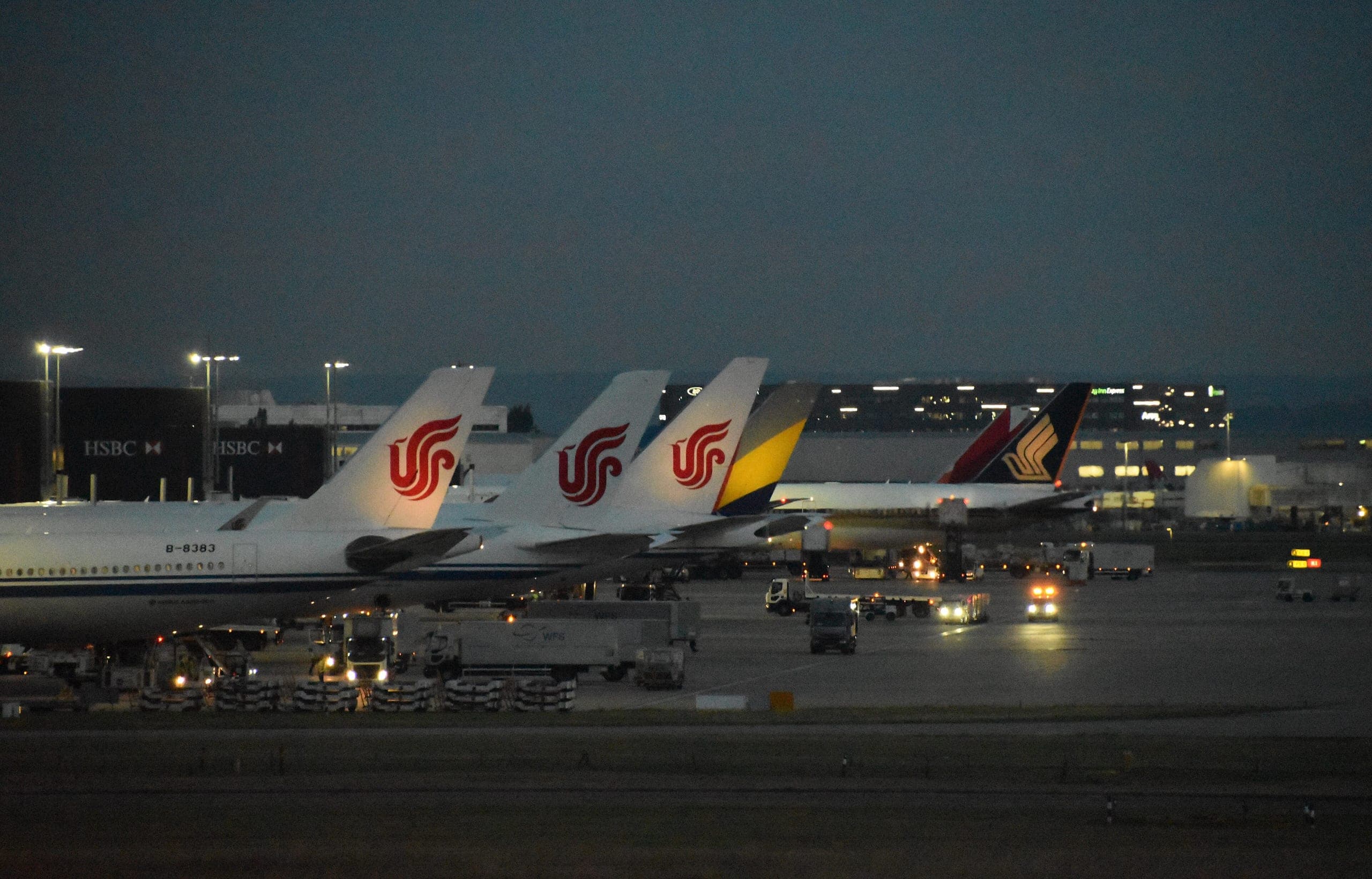 Air China and Singapore Airlines tails on the Terminal 2 apron at dusk, ground vehicles working beneath the floodlights