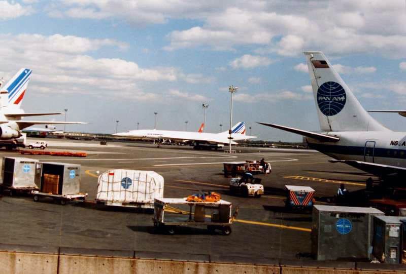 An Air France Concorde on the ramp at JFK around 1987, beside the tail of a Pan Am Boeing 747 and airport ground equipment