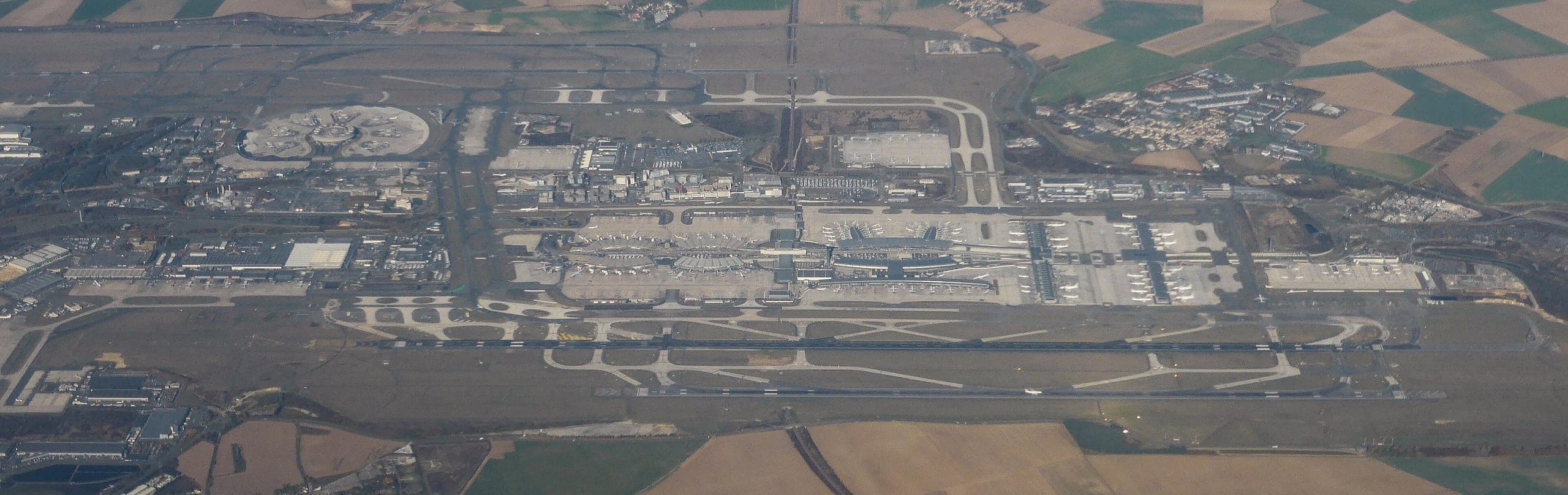 A wide aerial panorama across the whole airport, terminals in the centre between the northern and southern runway doublets