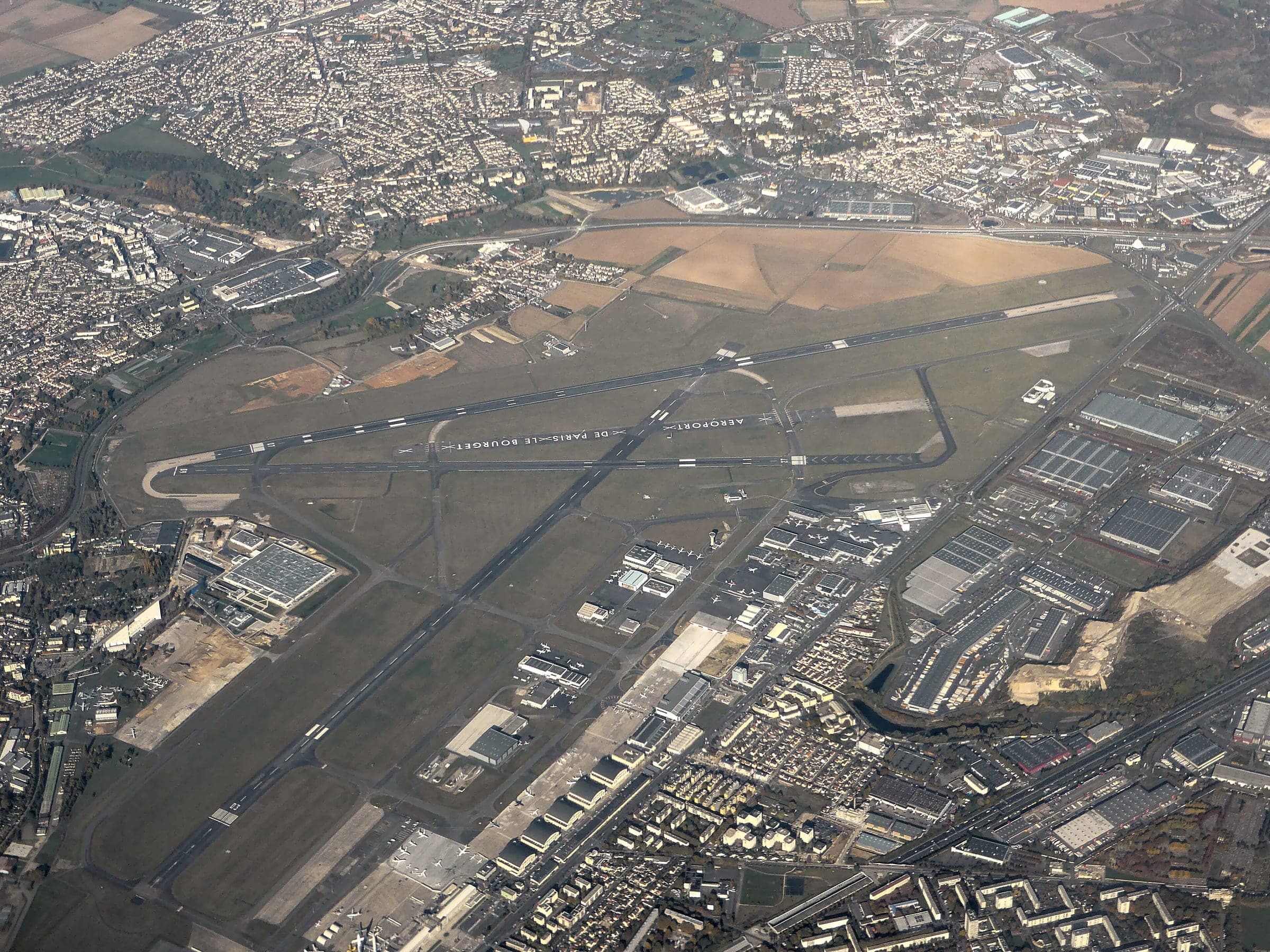 A steep aerial view of Le Bourget in autumn, looking along the runways with the museum aprons, business-aviation hangars and dense housing crowding the airfield boundary