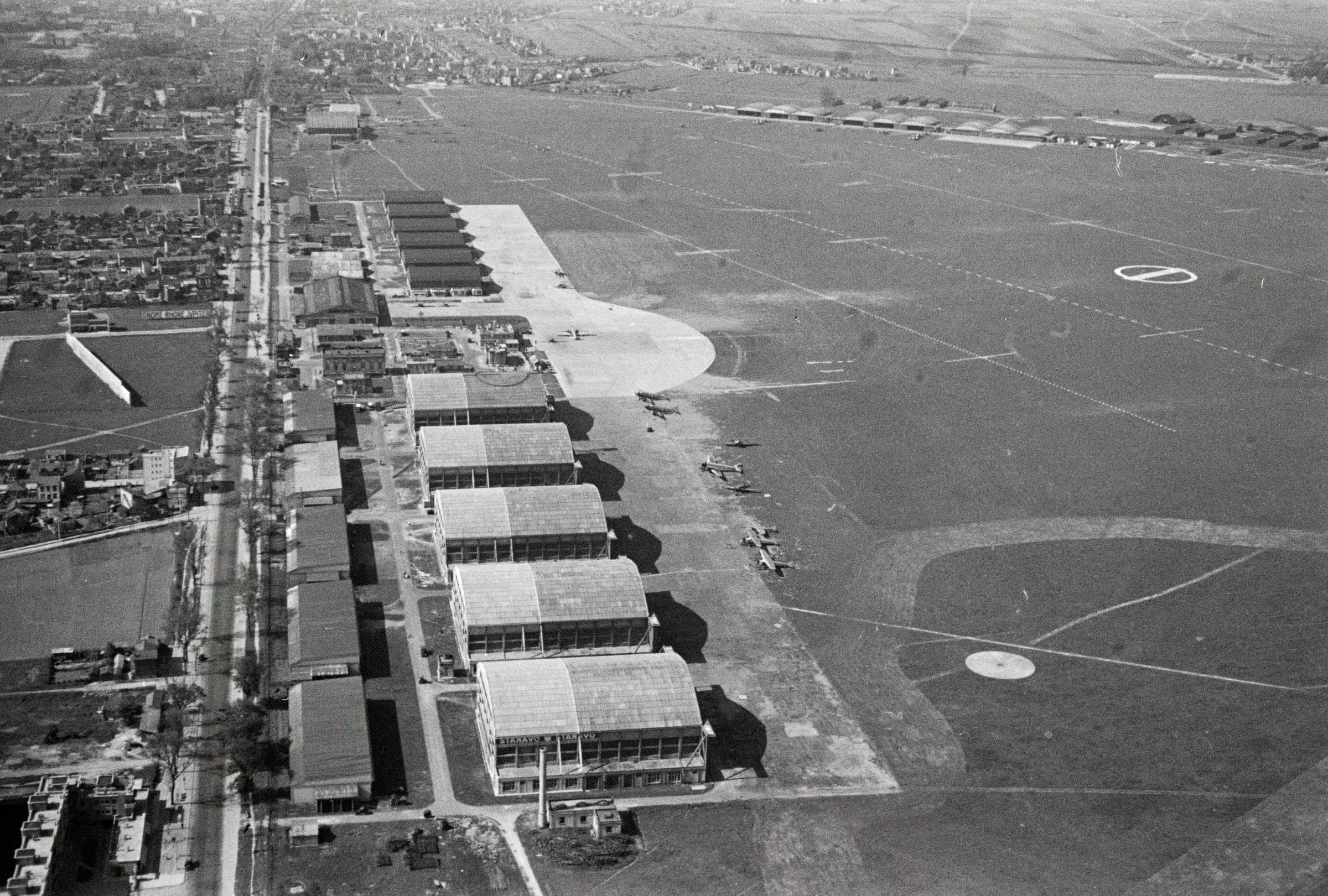 Oblique aerial view of Le Bourget in the early 1930s: a long row of arched hangars beside the road, the grass landing ground and its circle marker, before the new terminal was built