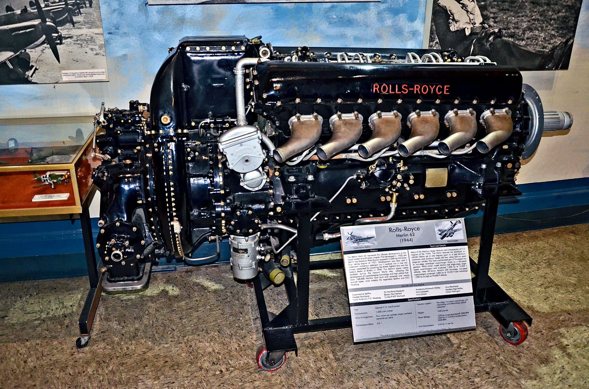 A 1944 Rolls-Royce Merlin 62 in side profile on a museum stand, its black cylinder blocks, exhaust stubs and reduction gear housing clearly lit