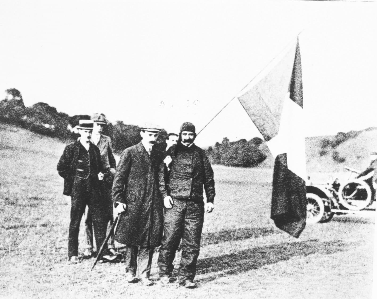 Three men in coats and caps standing on a grassy slope beside a flag and a motor car, catalogued as Blériot after the Channel flight.