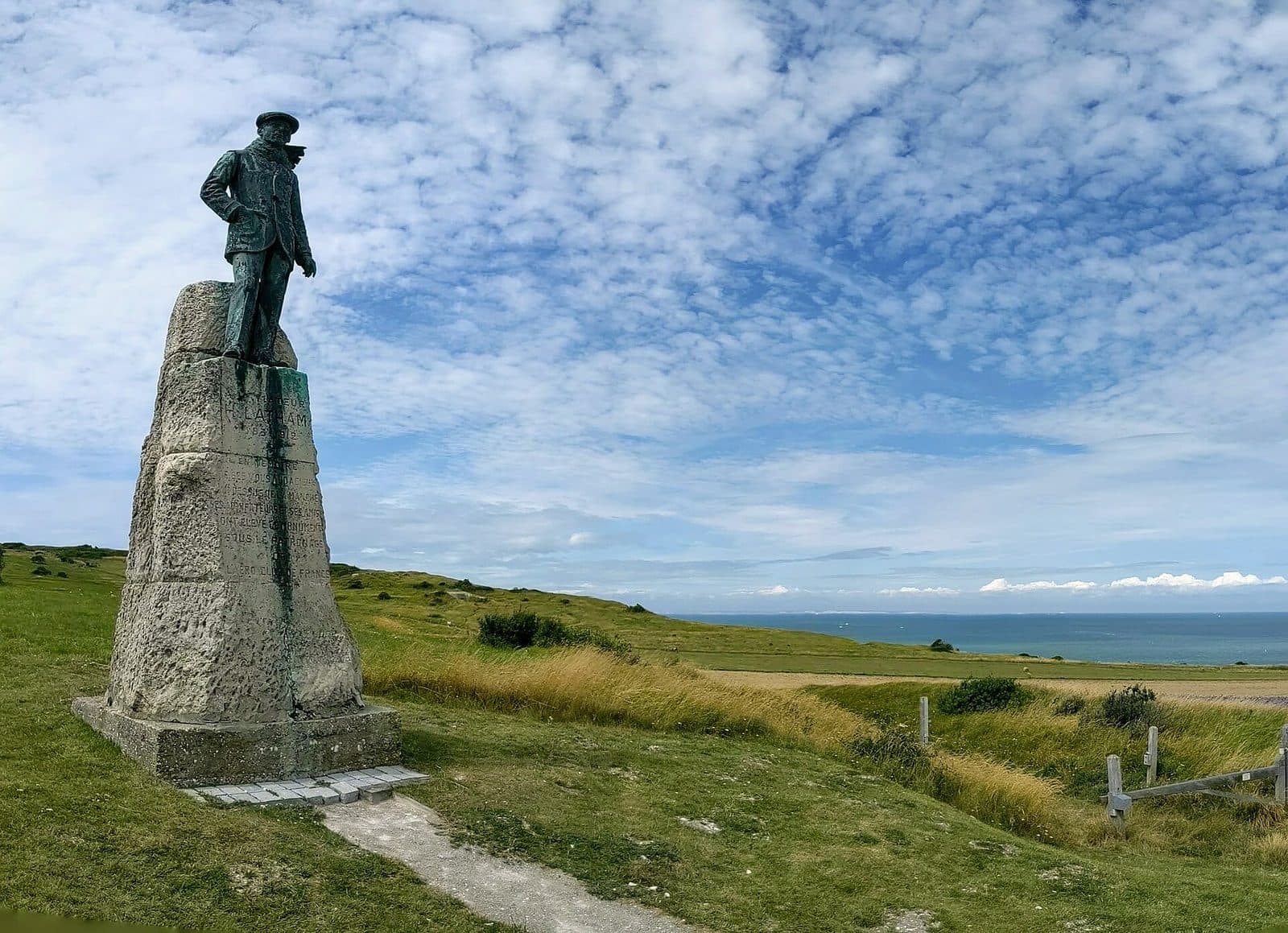 The statue of Hubert Latham on a stone plinth on the clifftop grassland near Sangatte, the Channel behind.