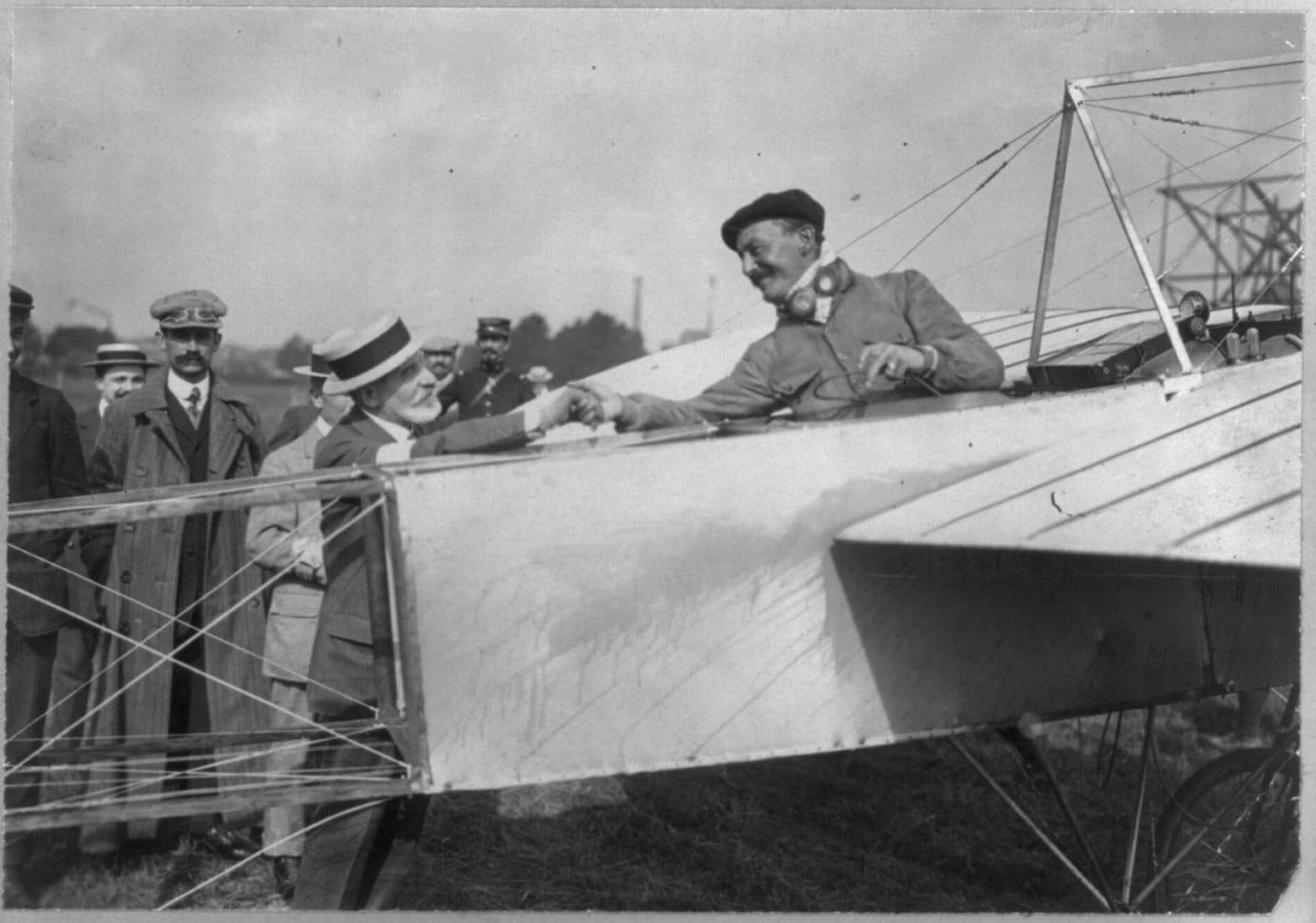 Alfred Leblanc, in cap and coat, leaning over the side of a Blériot monoplane among a crowd.