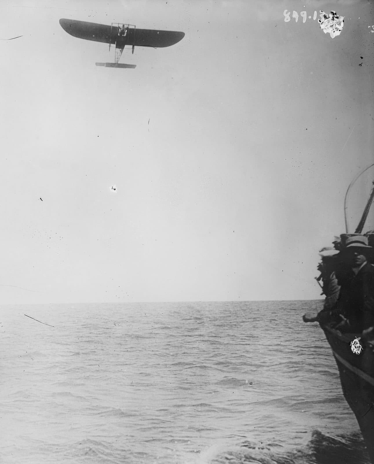 A tiny monoplane high over open water, the rail of a ship at the edge of the frame, catalogued as Blériot in mid-Channel.