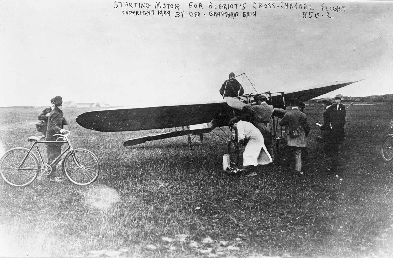 The Blériot XI on the grass at Les Baraques, men bent over its engine and a cyclist watching, as the motor is started for the cross-Channel flight.