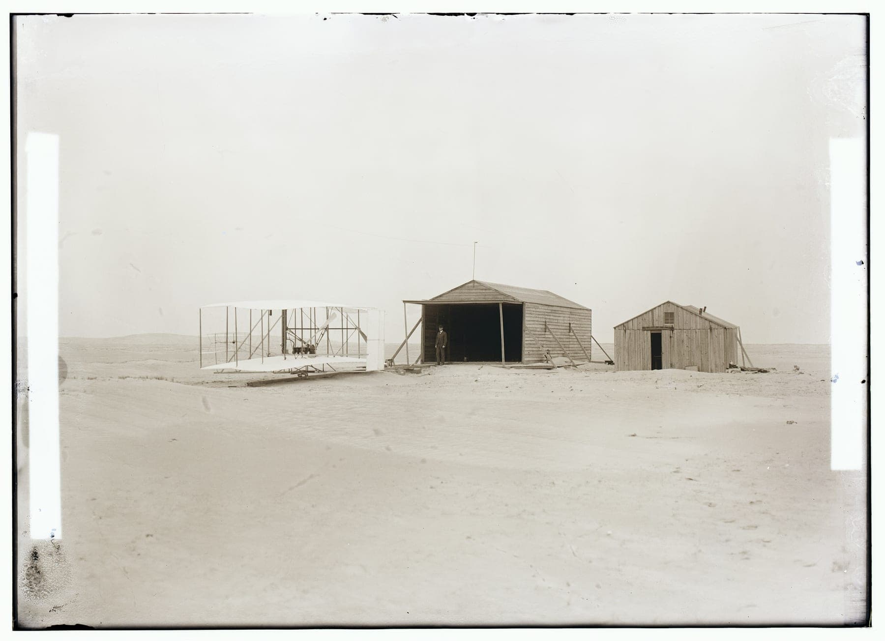 The 1903 Flyer outside the camp buildings at Kill Devil Hills: a large shed housing the machine and a smaller hut used as workshop and living quarters.
