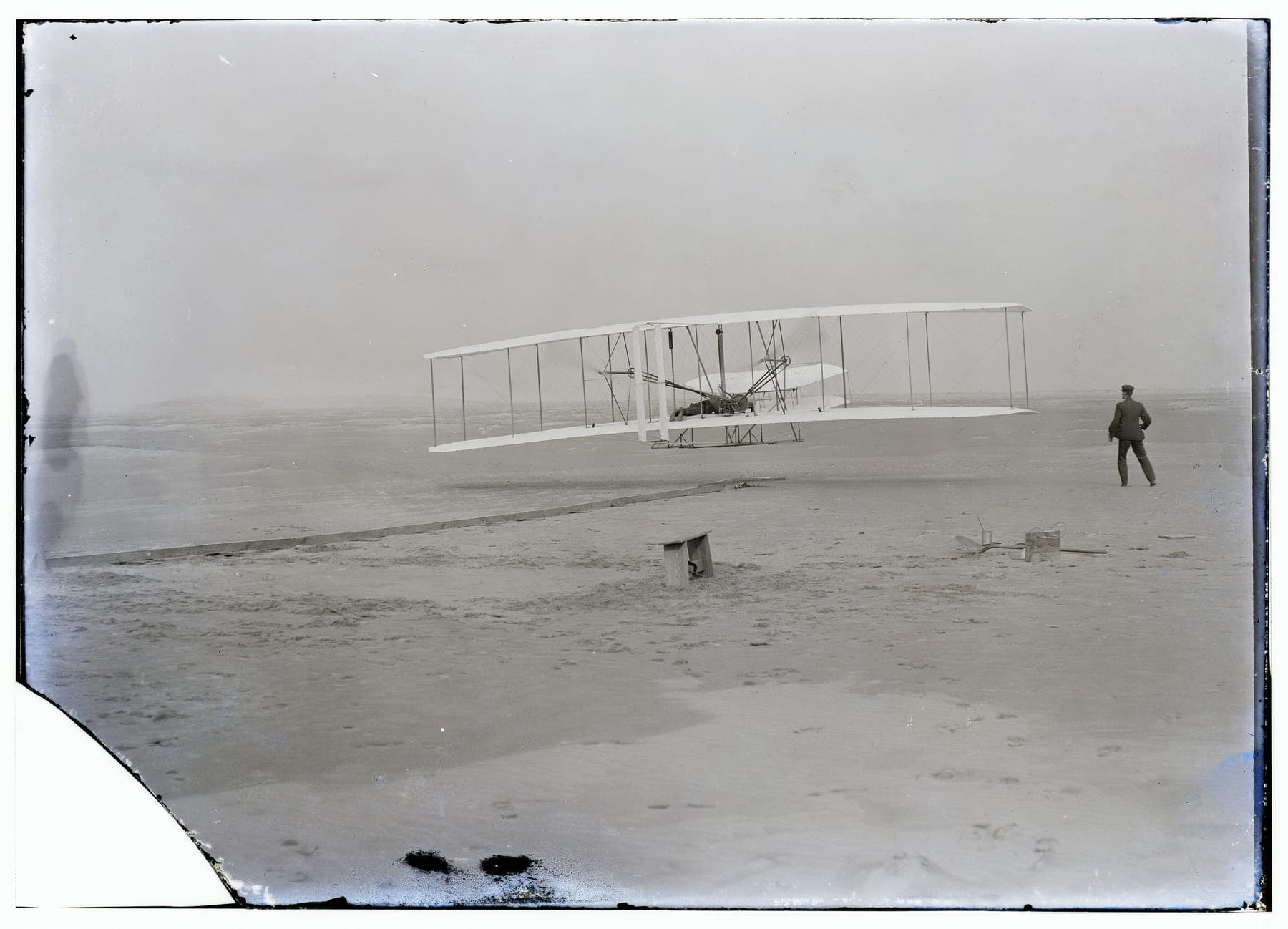 The Wright Flyer just airborne over the sand at Kill Devil Hills, Orville lying prone at the controls and Wilbur running at the wingtip.
