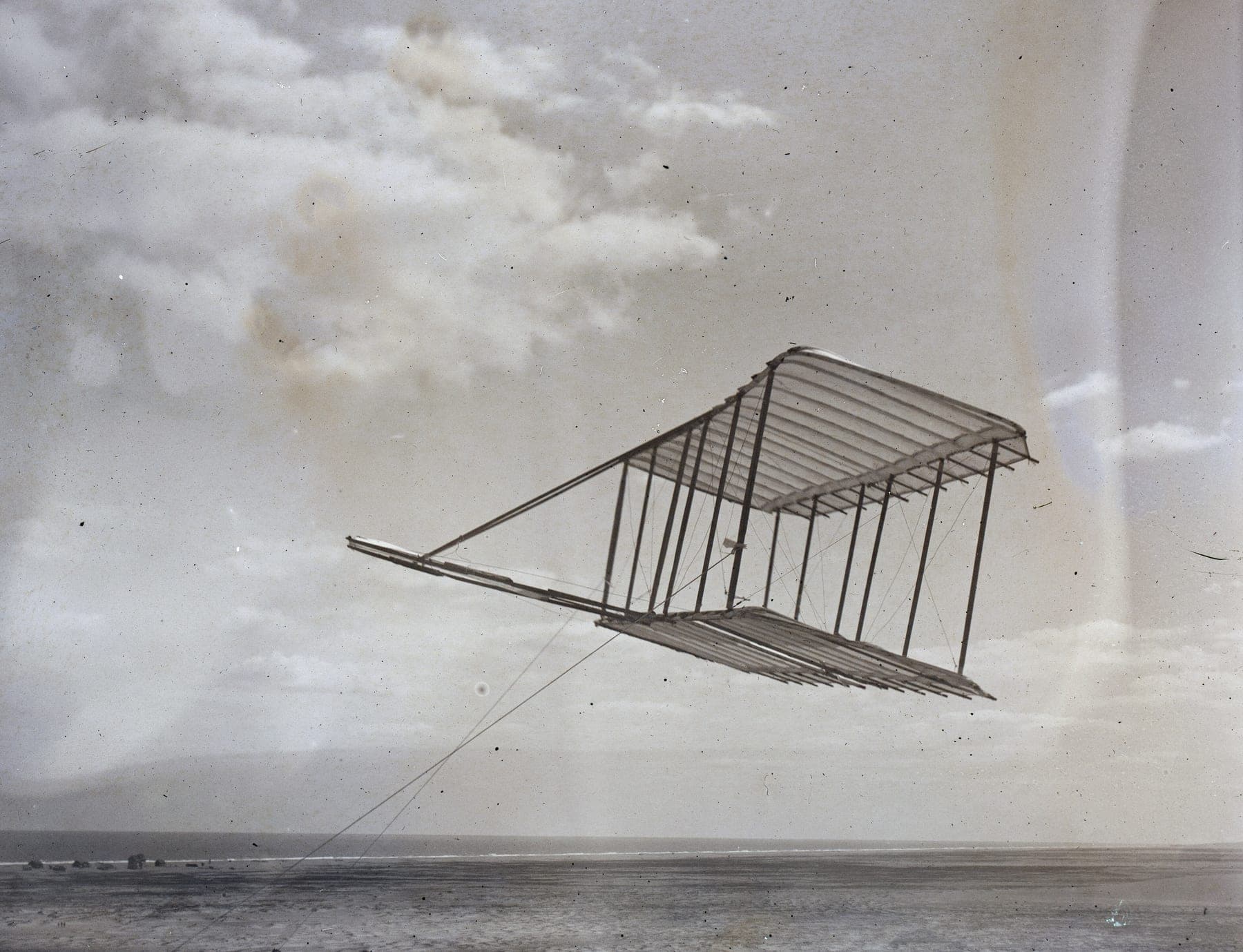 The 1900 glider flying as a kite on its lines above the sand at Kitty Hawk, its forward elevator clearly visible.
