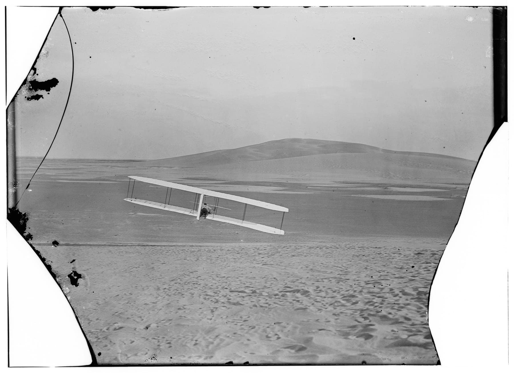 The 1902 glider in a right turn, seen from behind, its right wingtip close to the ground.