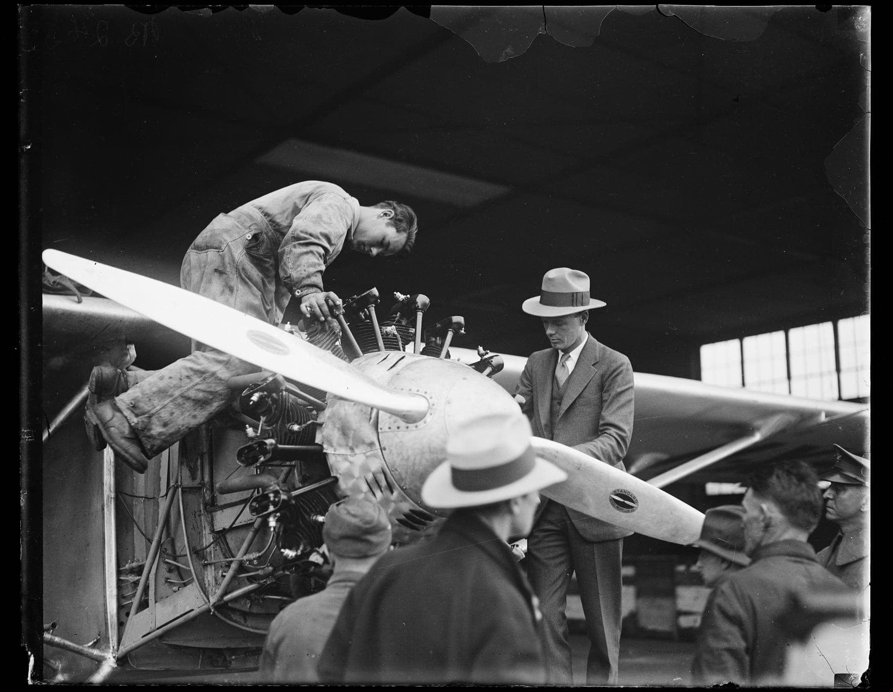 Lindbergh, in a suit and hat, inspecting the Spirit's Whirlwind engine while a mechanic works on the cylinders.