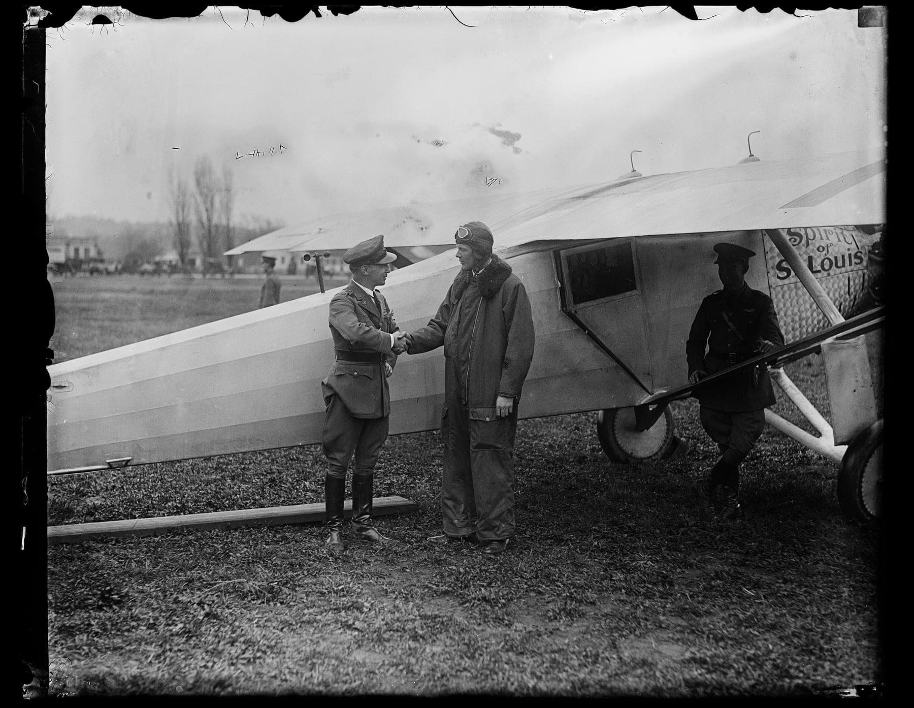 Charles Lindbergh, in flying suit and helmet, shaking hands with an Army officer beside the Spirit of St. Louis, its name painted on the engine cowling.